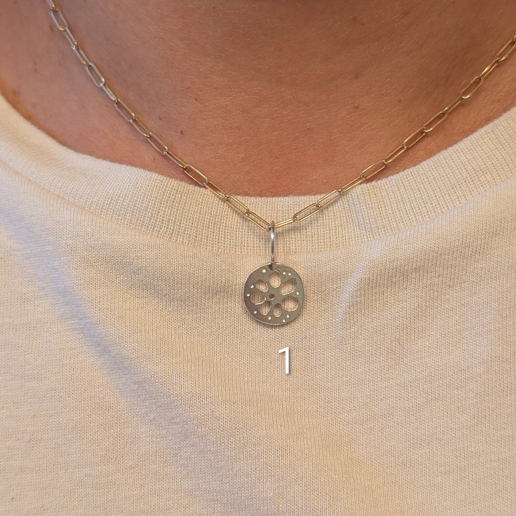 A close-up of a person's neck and chest wearing a delicate gold chain necklace with a circular silver-toned pendant featuring a floral or wheel-like cutout pattern. The person is wearing a cream-colored graphic t-shirt.