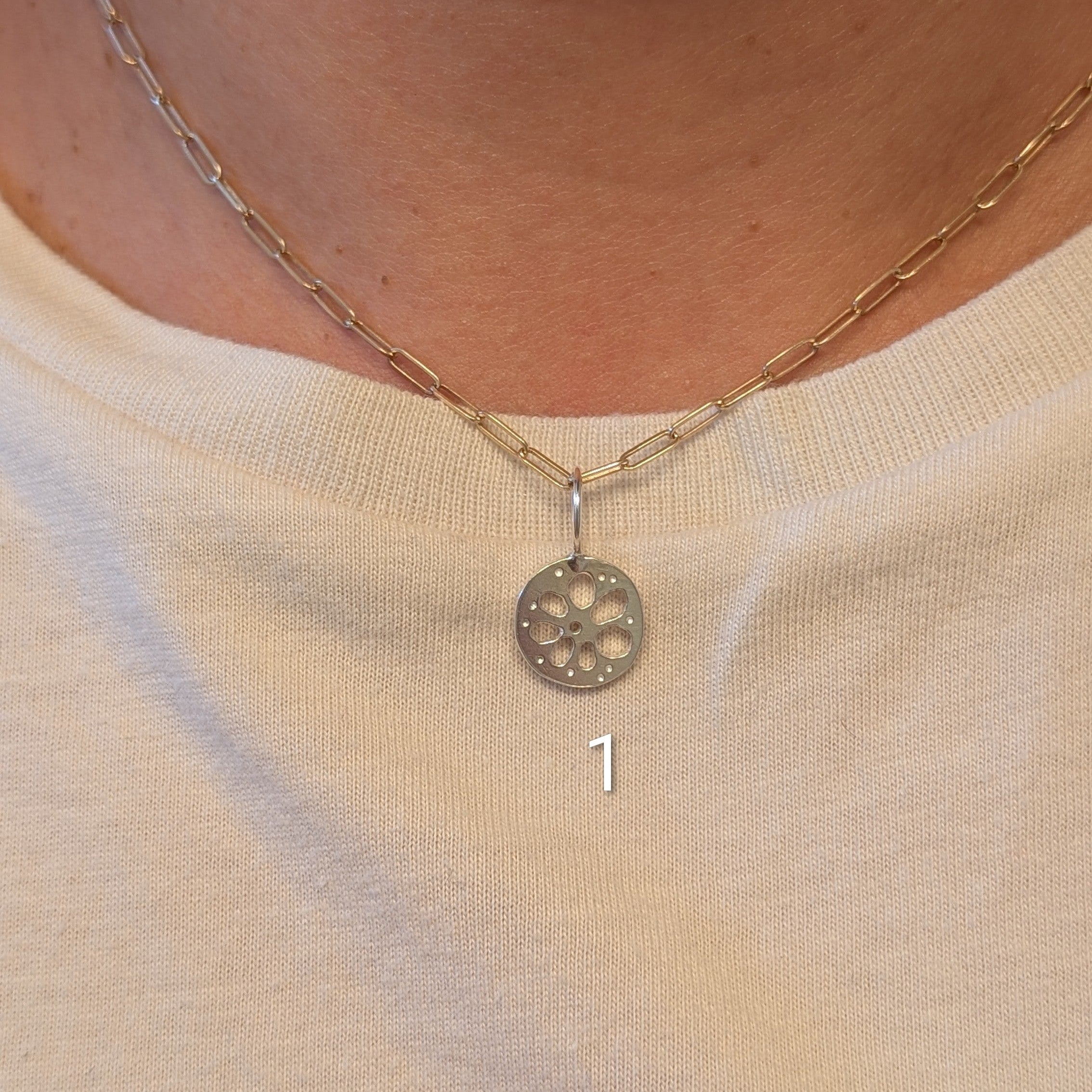 A close-up of a person's neck and chest wearing a delicate gold chain necklace with a circular silver-toned pendant featuring a floral or wheel-like cutout pattern. The person is wearing a cream-colored graphic t-shirt.