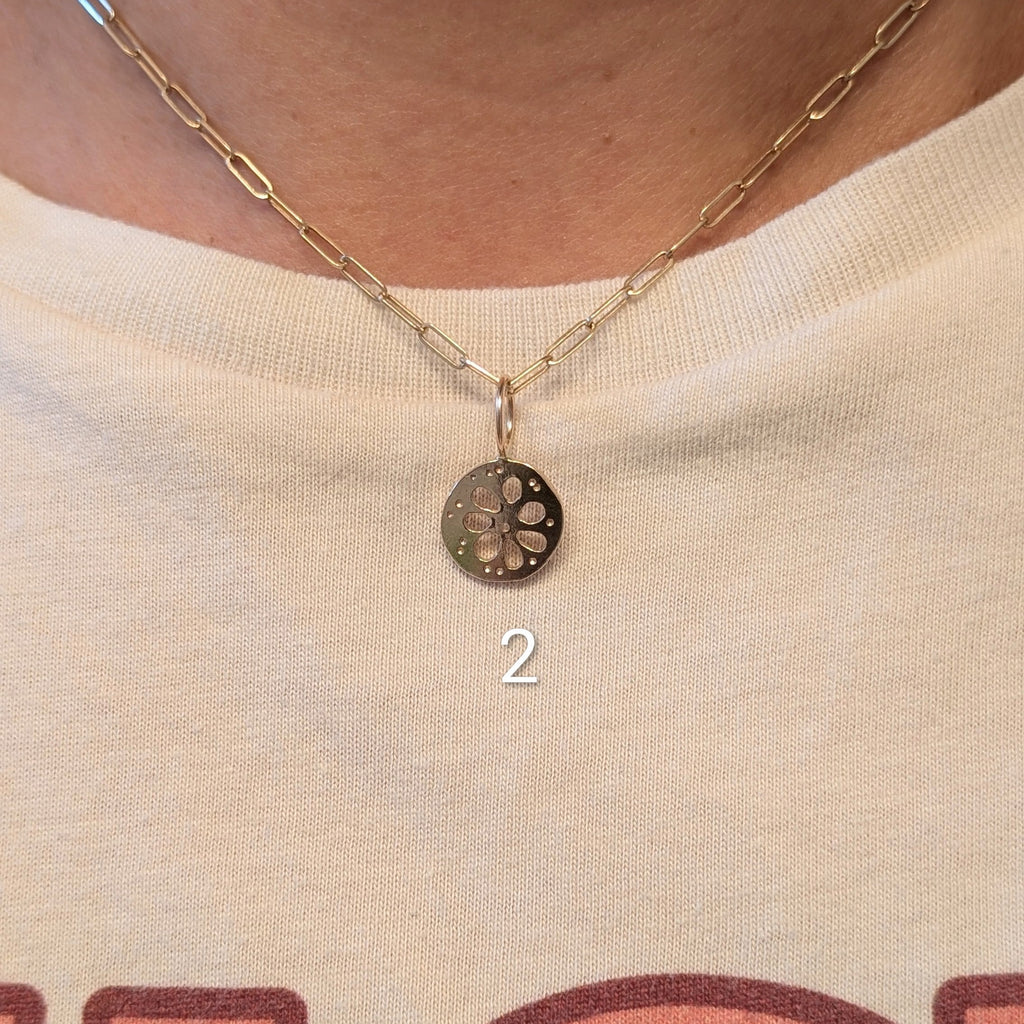 A close-up of a person wearing a delicate gold chain necklace with a small circular pendant featuring a lotus root design. The necklace is shown on the neck over a cream-colored graphic t-shirt.