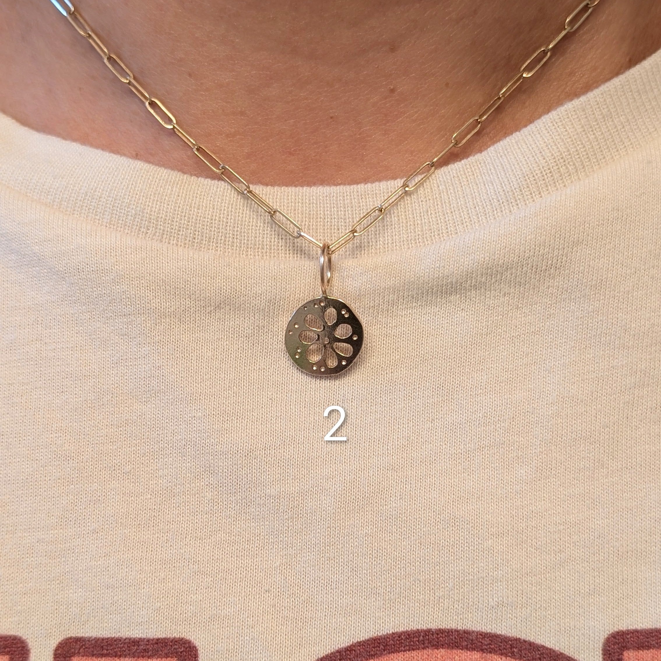 A close-up of a person wearing a delicate gold chain necklace with a small circular pendant featuring a lotus root design. The necklace is shown on the neck over a cream-colored graphic t-shirt.