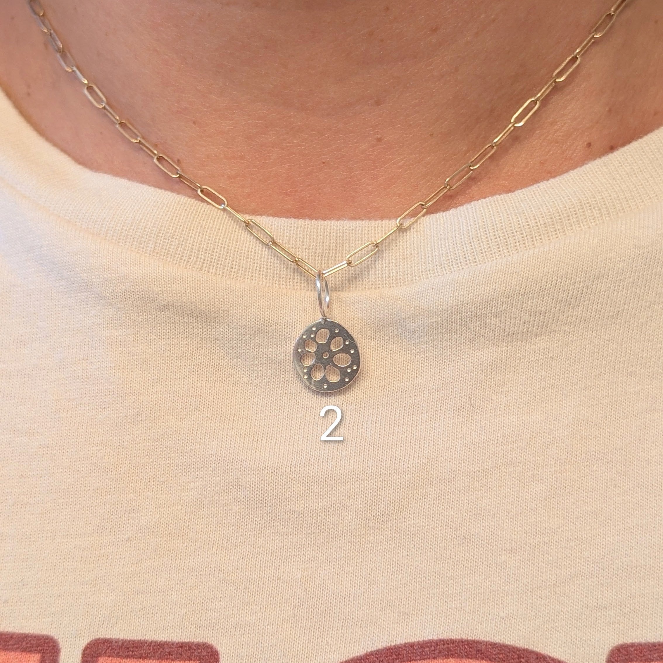 A close-up of a person wearing a delicate gold chain necklace with a small circular pendant featuring a lotus root design. The necklace is shown on the neck over a cream-colored graphic t-shirt.
