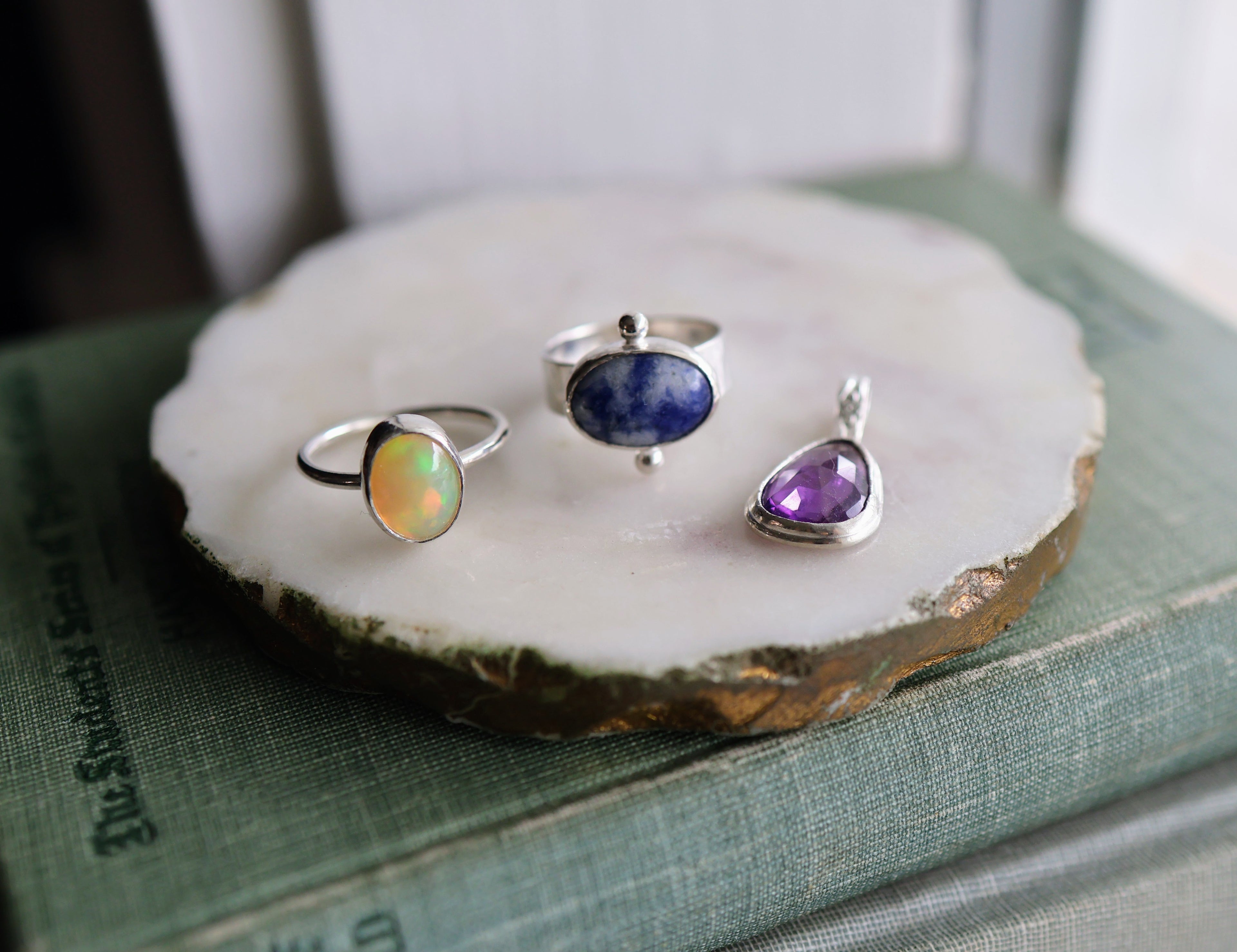 An opal and silver ring, a blue jasper and silver ring, and an amethyst pendant on a white display dish, on top of a stack of vintage books.
