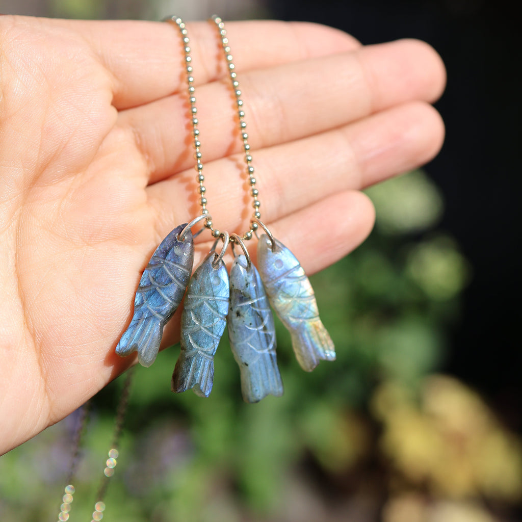 Hand holding necklace with 4 labradorite fish pendants with a blurred natural background