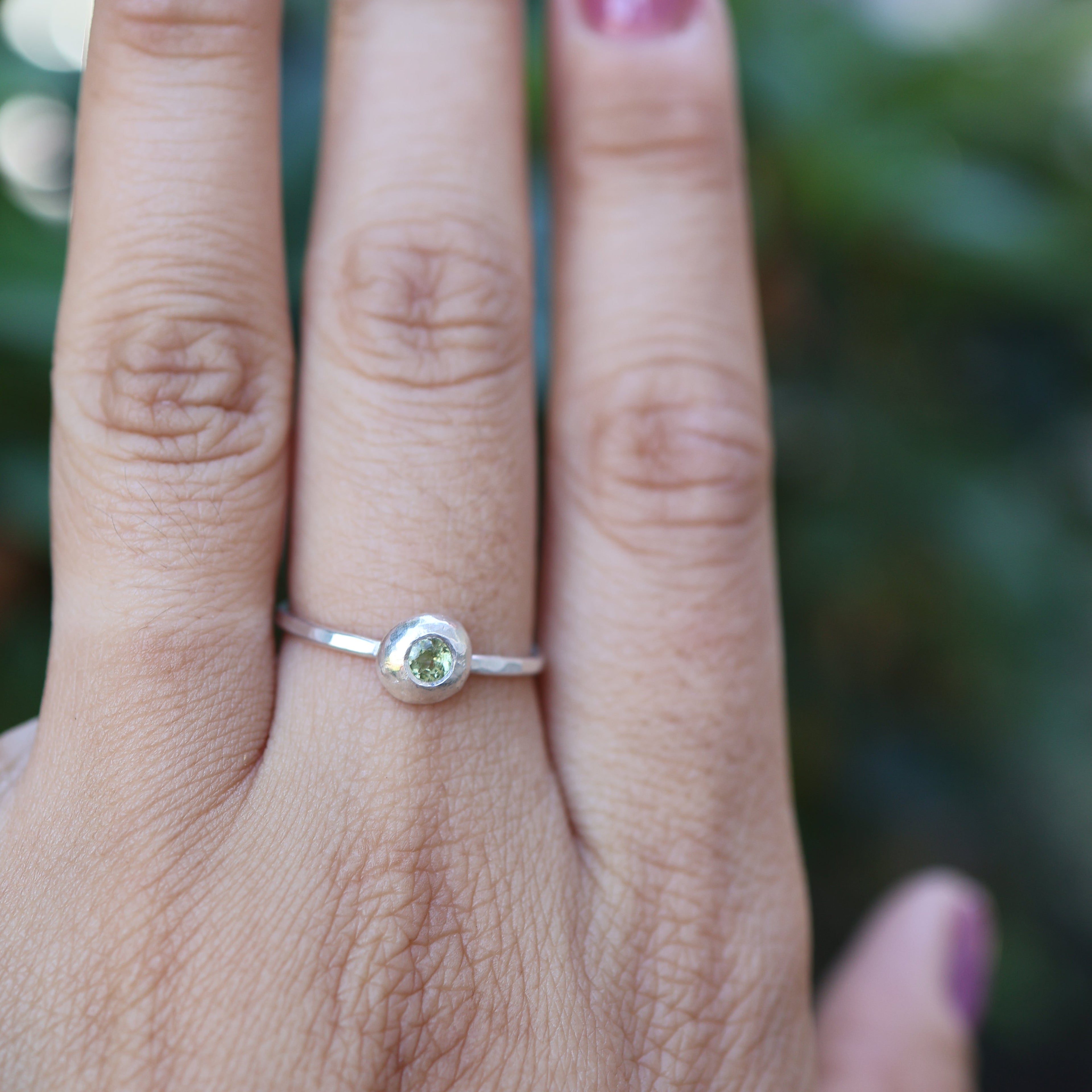 Hand wearing a silver ring with a green gemstone against a blurred natural background