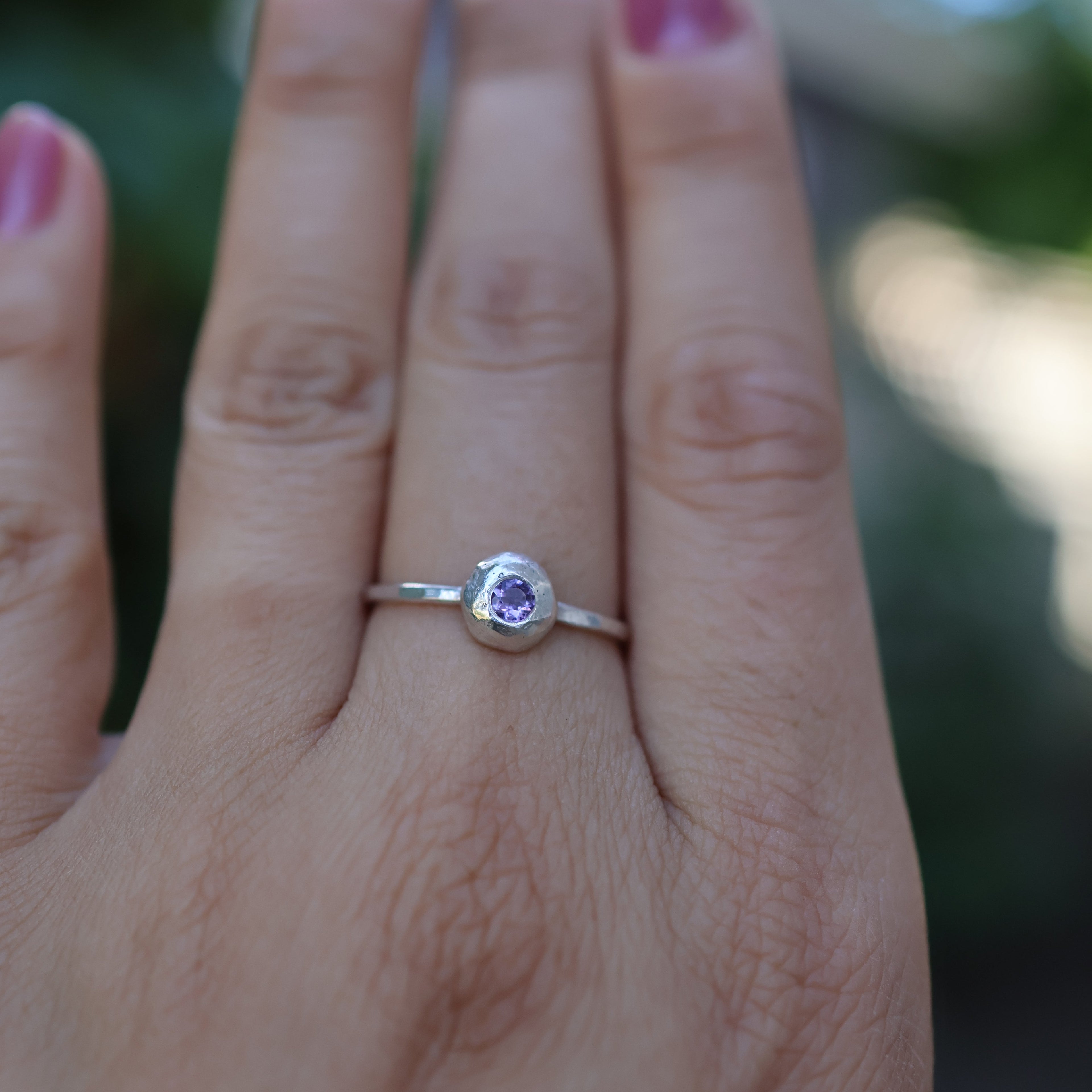 Hand wearing a silver ring with a purple gemstone against a blurred natural background