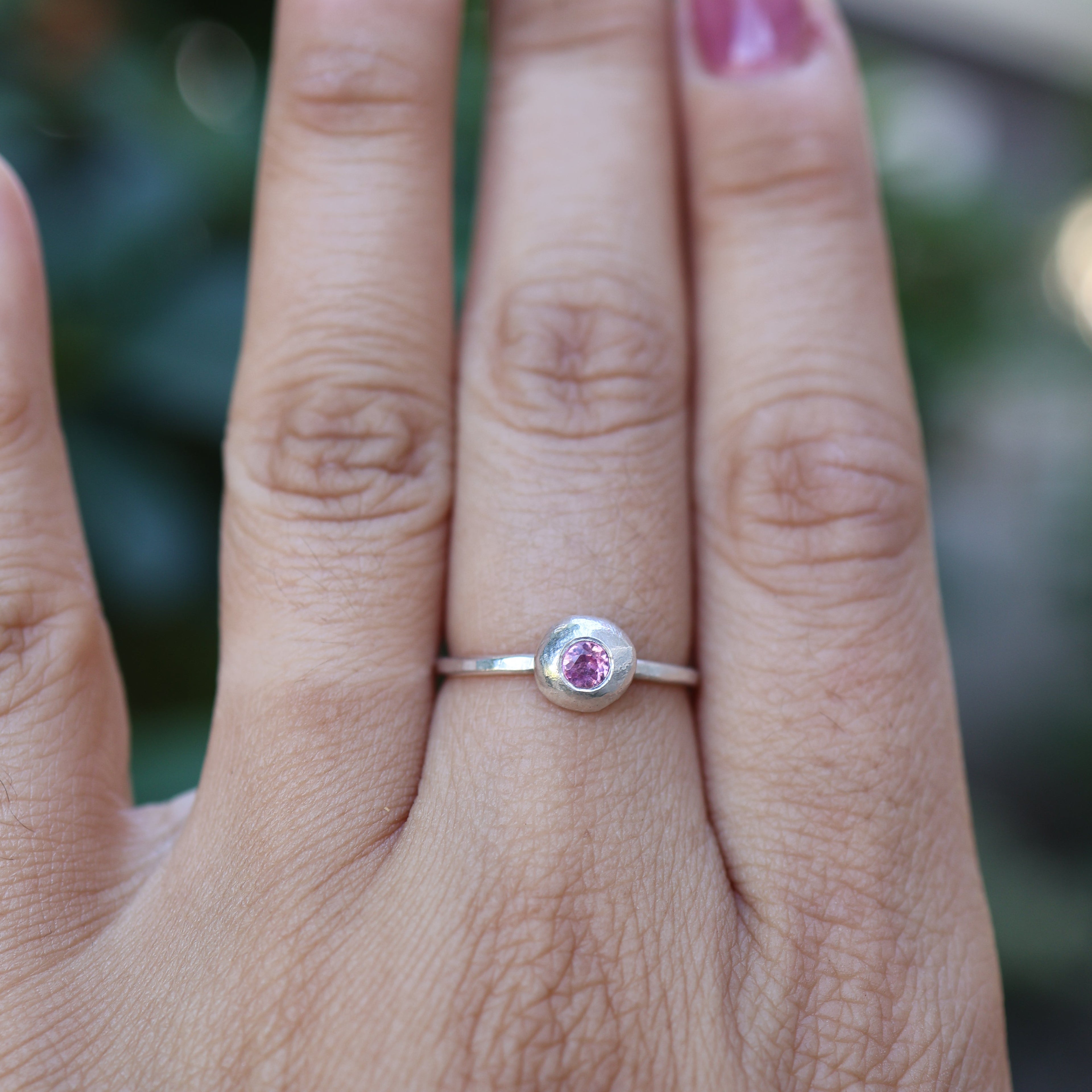Hand wearing a silver ring with a pink gemstone against a blurred green background