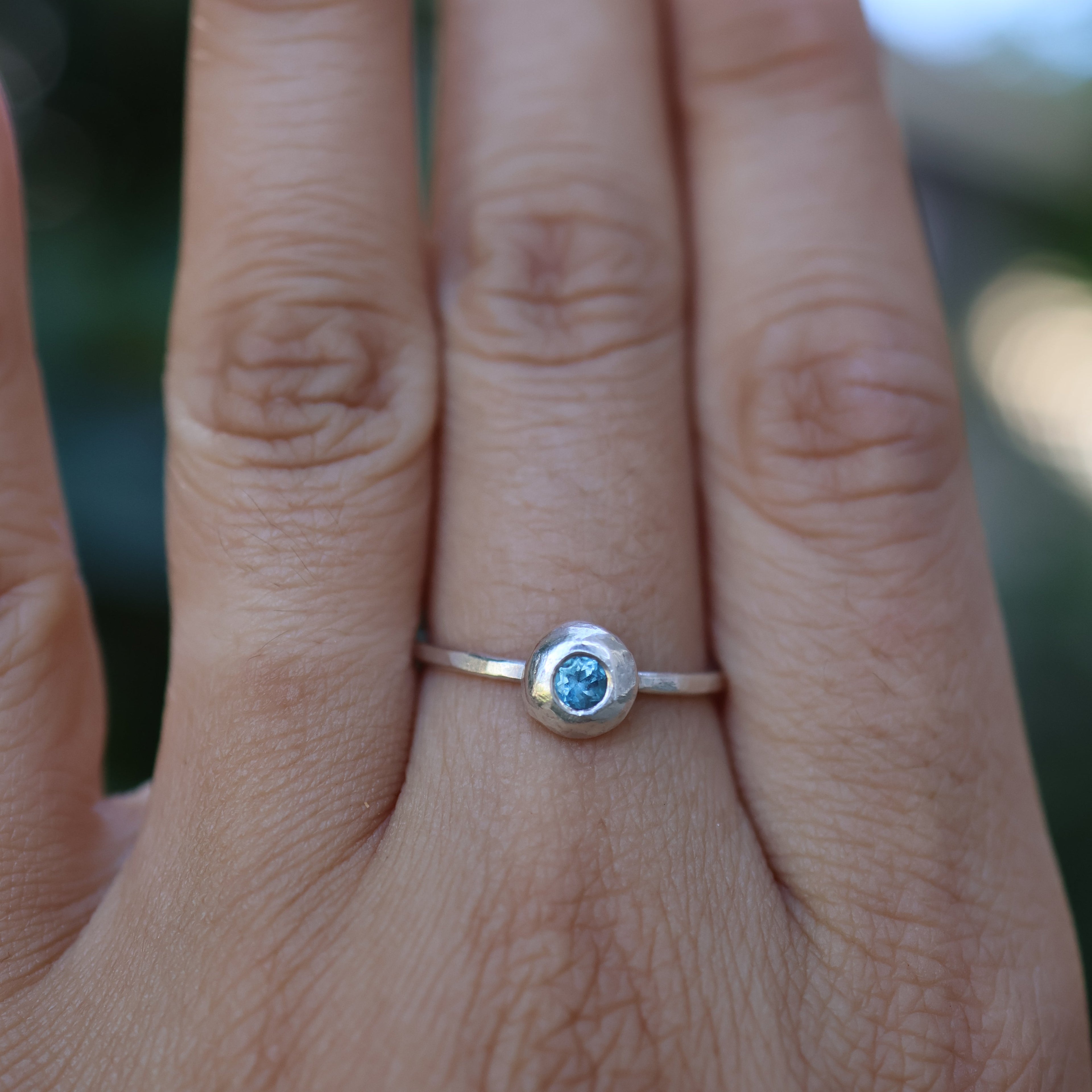 Close-up of a hand wearing a silver ring with a blue gemstone against a blurred background
