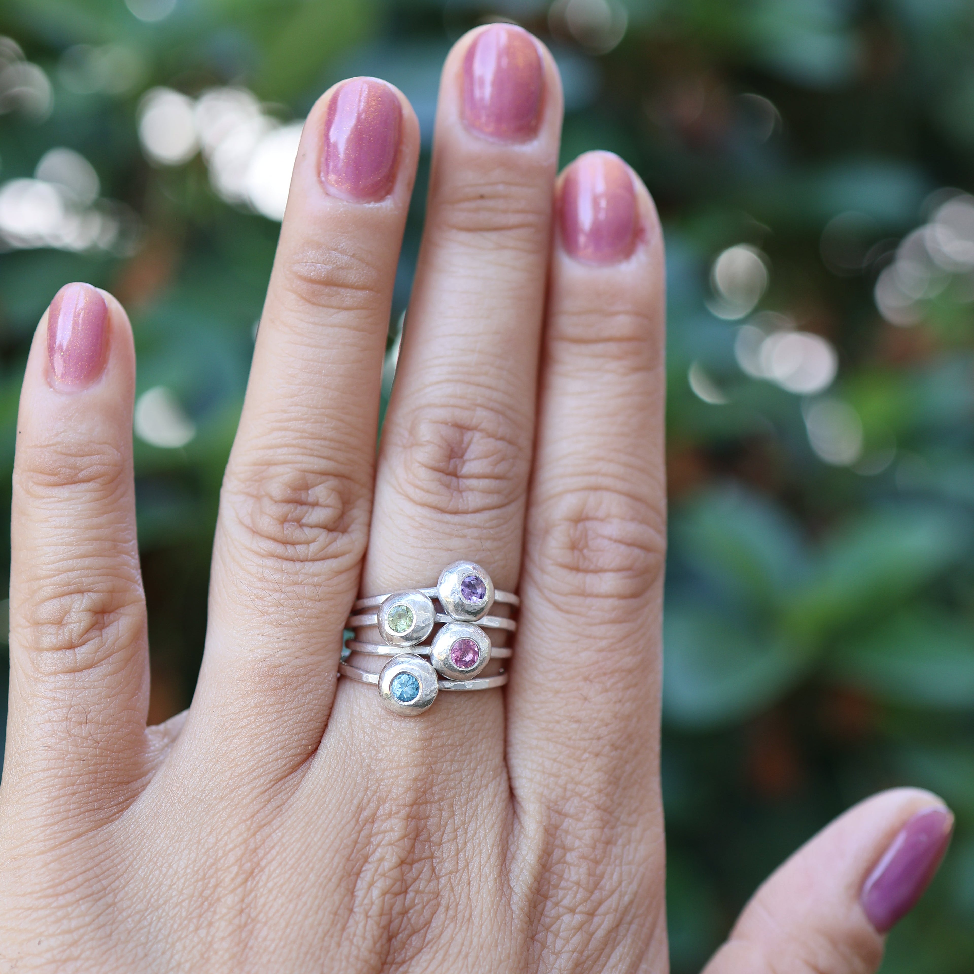 Hand wearing a stack of rings with colorful gemstones against a blurred green background