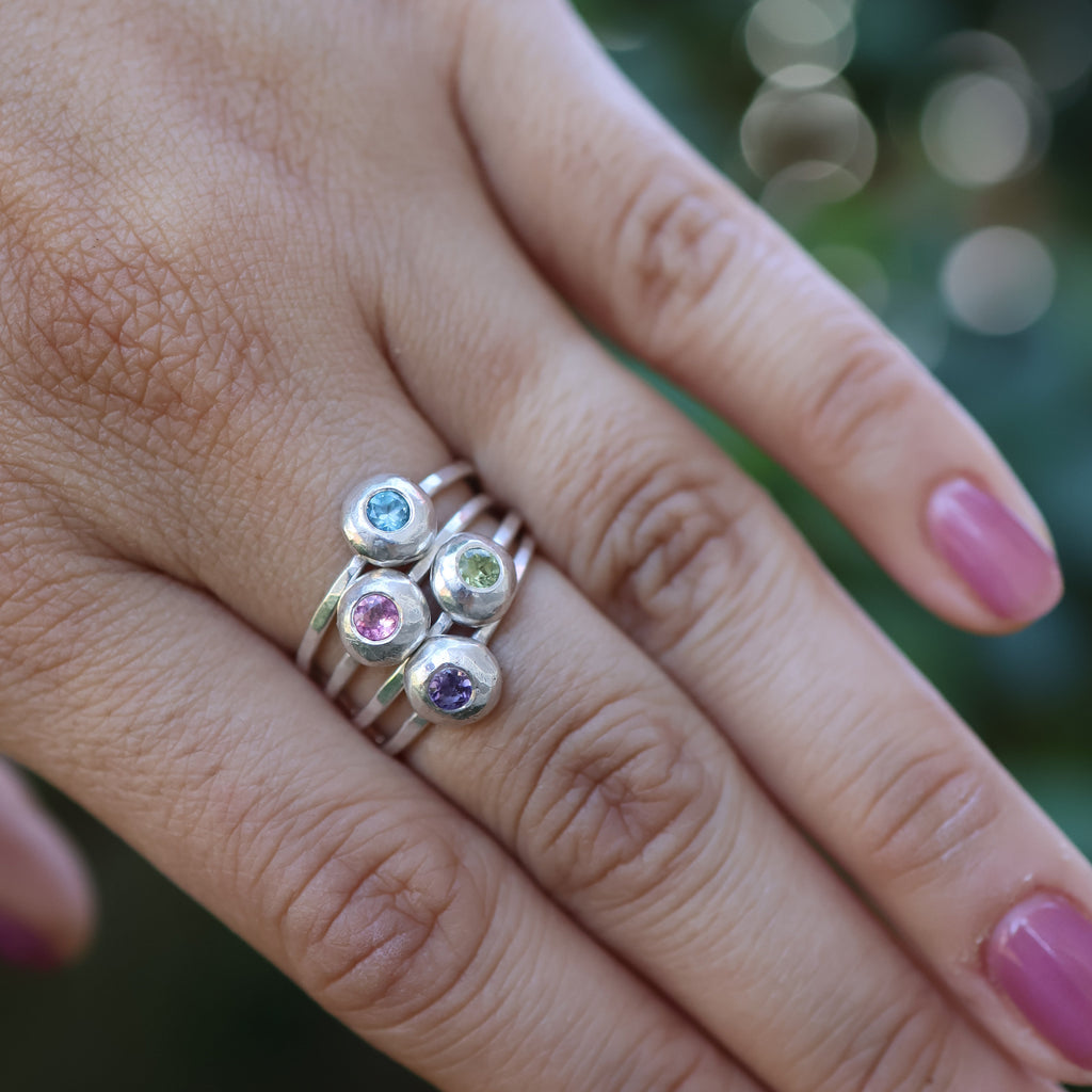 Hand wearing a stack of silver rings with multicolored gemstones against a blurred natural background