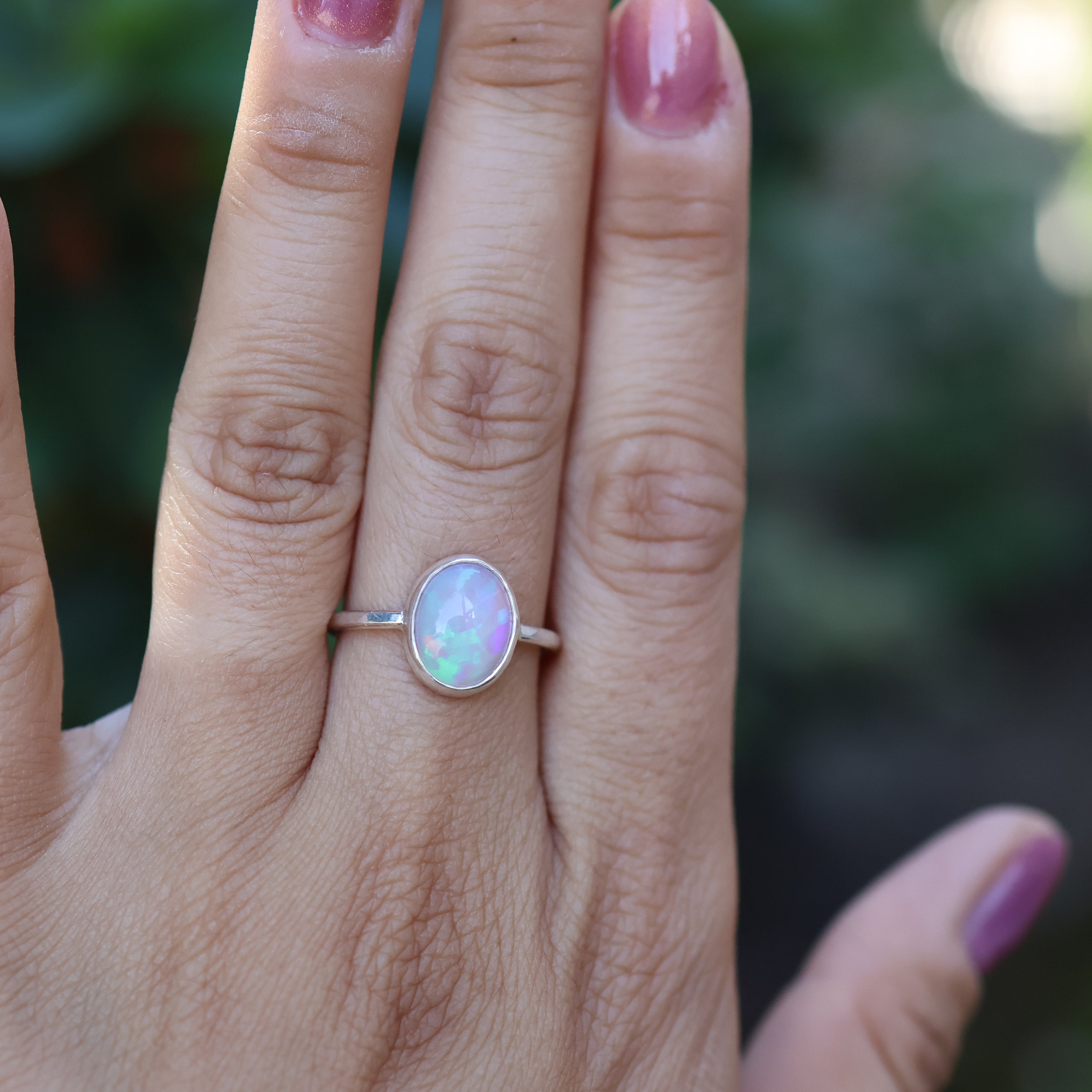 Hand wearing a silver ring with an opal stone against a blurred green background