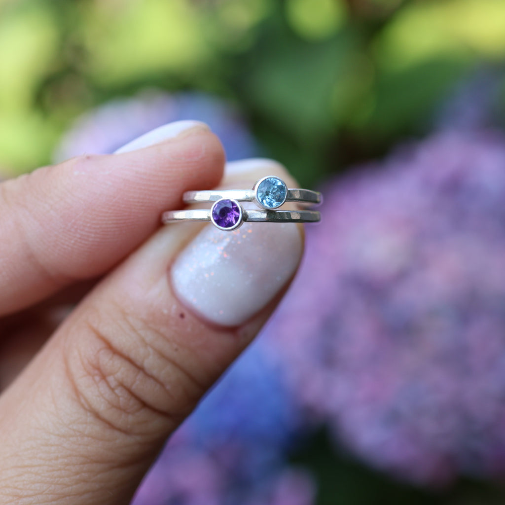 A close-up of a hand holding two thin silver rings with small round gemstones, one purple and one light blue, against a blurred background of green foliage and purple flowers.