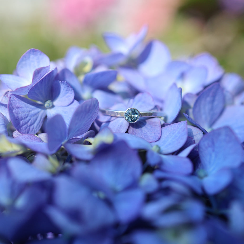 A delicate silver ring with a small round blue gemstone resting on a cluster of blue-purple hydrangea flowers.