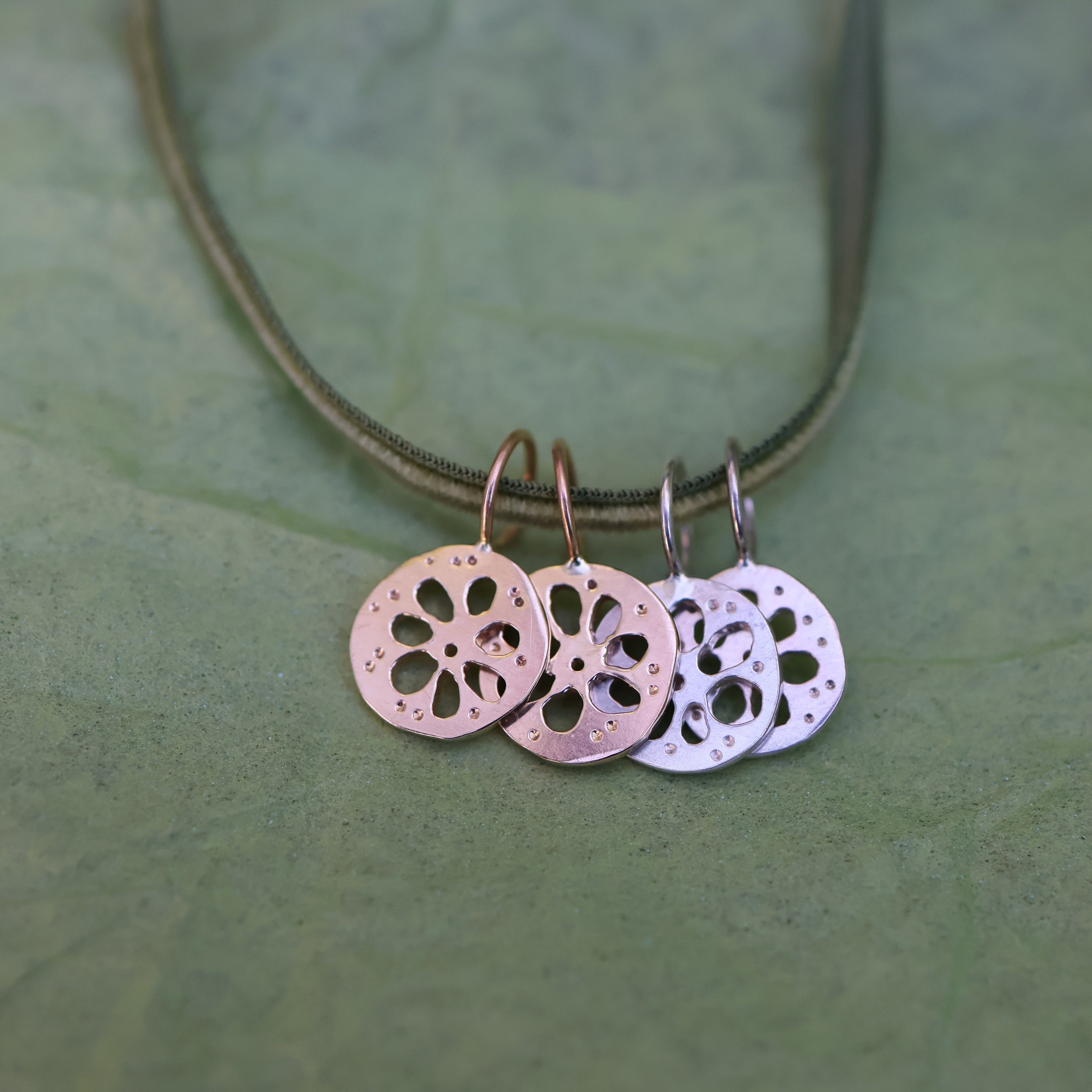 Four lotus root pendants, two gold and two silver, hanging from a thin green cord, displayed on a textured green surface.