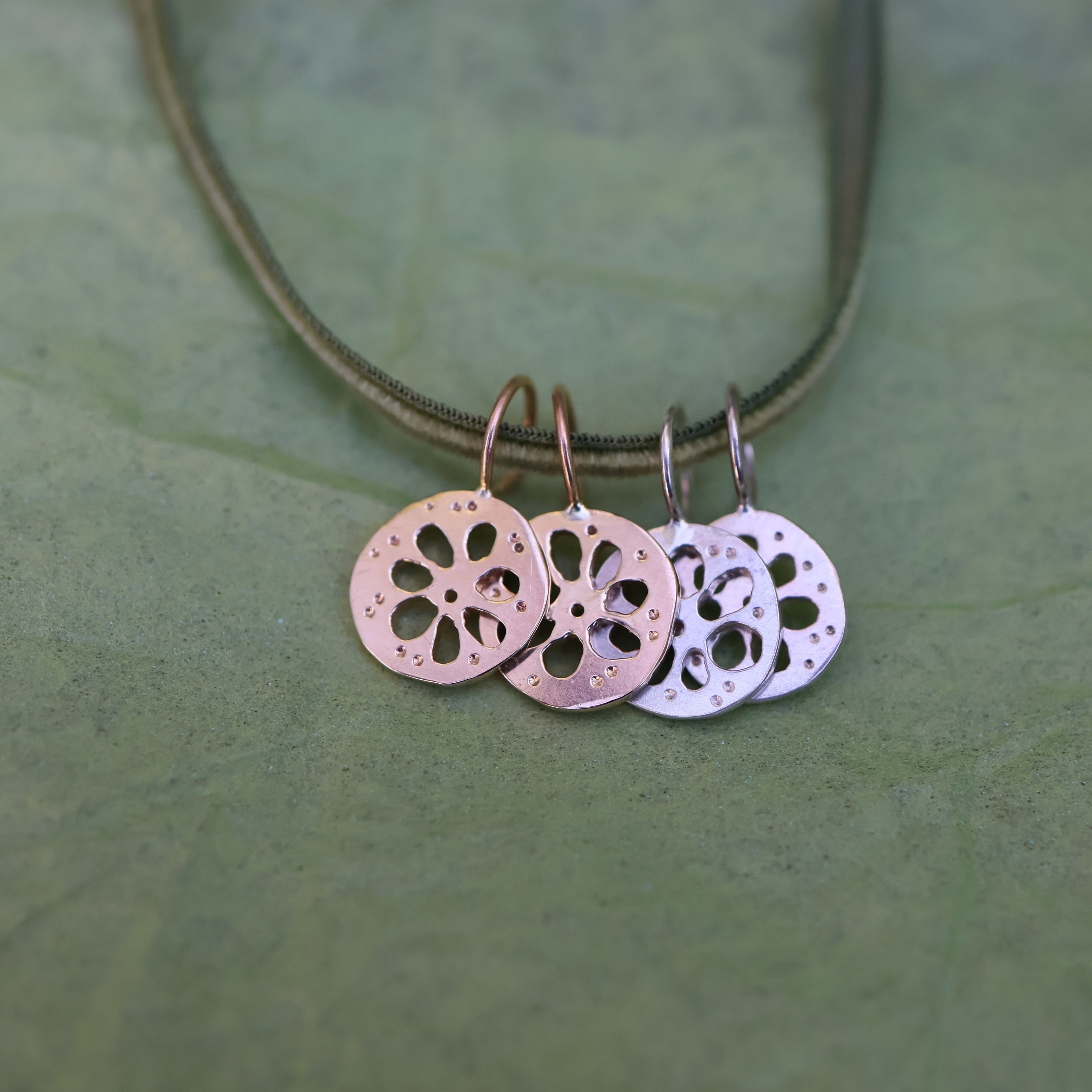 Four lotus root pendants, two gold and two silver, hanging from a thin green cord, displayed on a textured green surface.