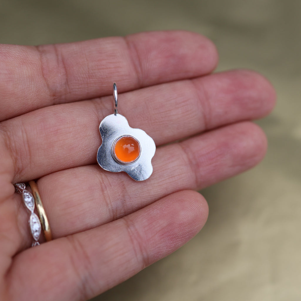 a hand holding a pendant in the shape of a sunny side up fried egg with a orange center stone