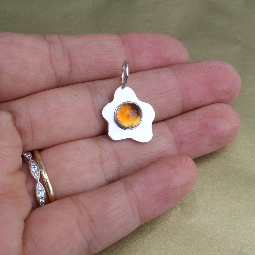a hand holding a pendant in the shape of a sunny side up fried egg with a orange center gemstone