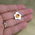 a hand holding a pendant in the shape of a sunny side up fried egg with a orange center gemstone
