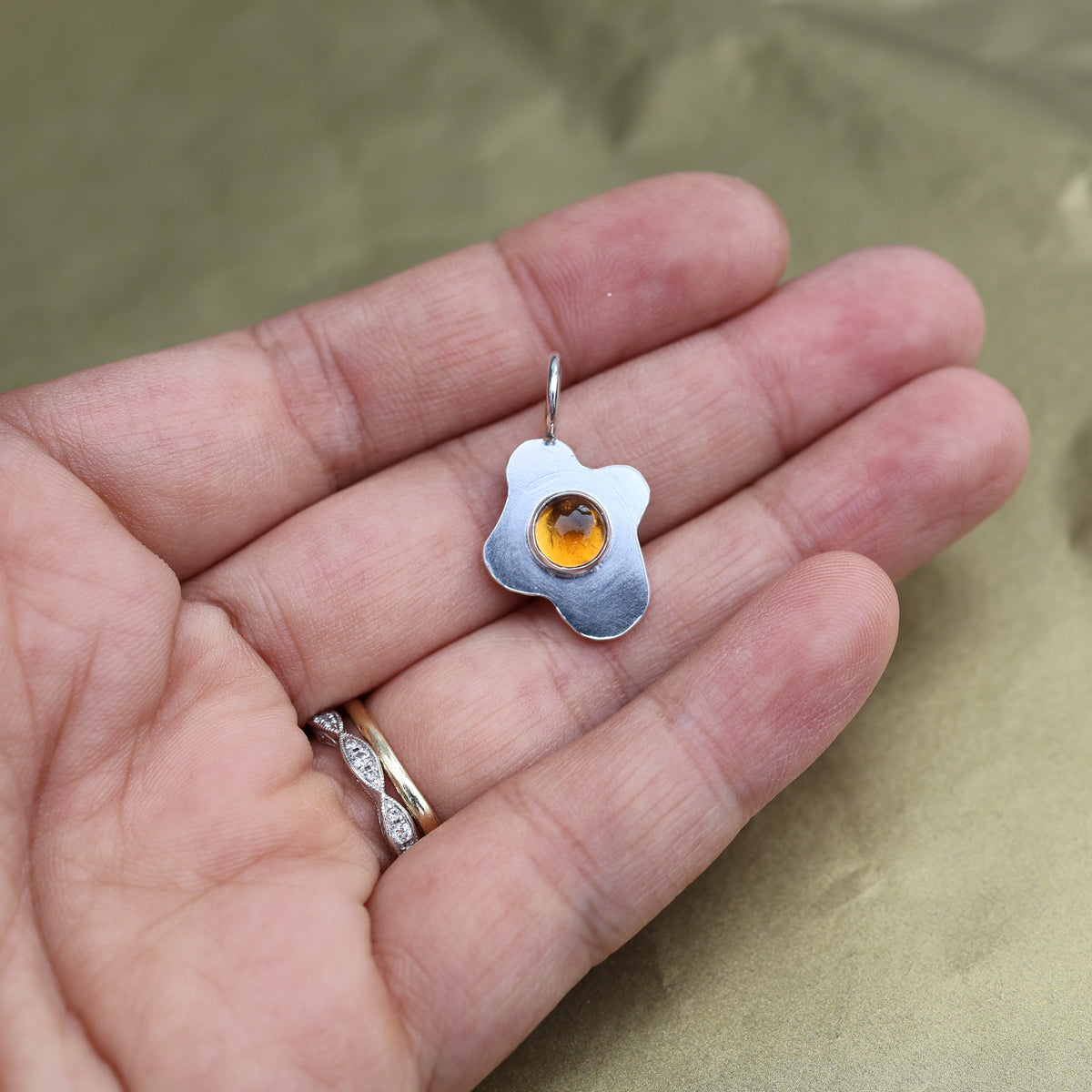 a hand holding a pendant in the shape of a sunny side up fried egg with a orange center gemstone
