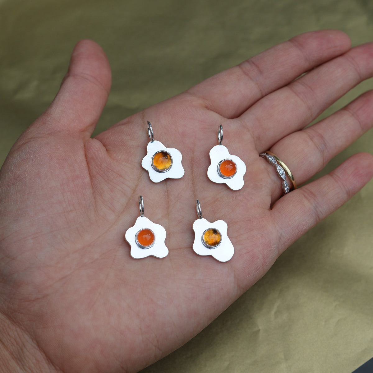 a hand holding four fried egg pendants with orange center gemstones