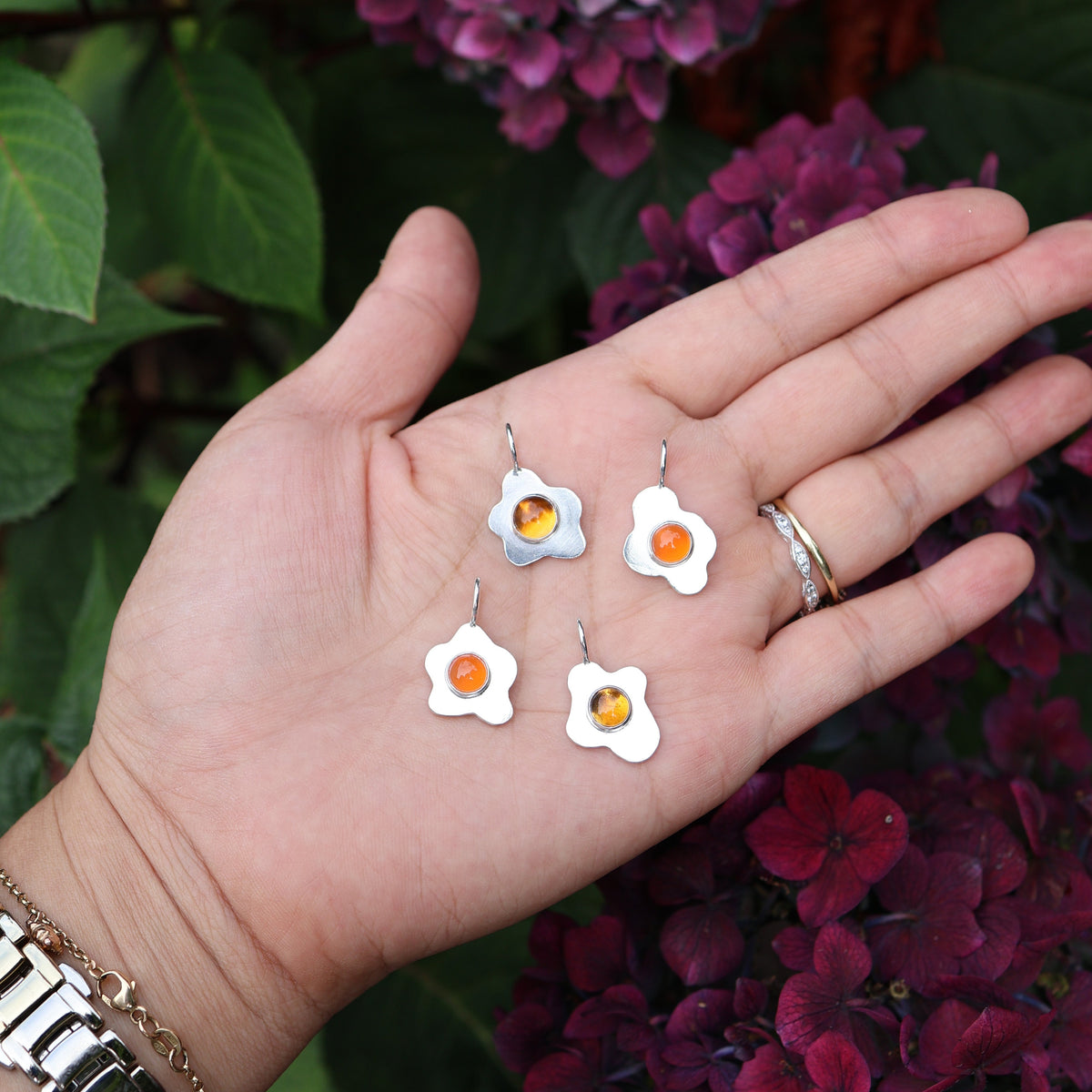 a hand holding four fried egg pendants with orange center gemstones, in front of a floral background