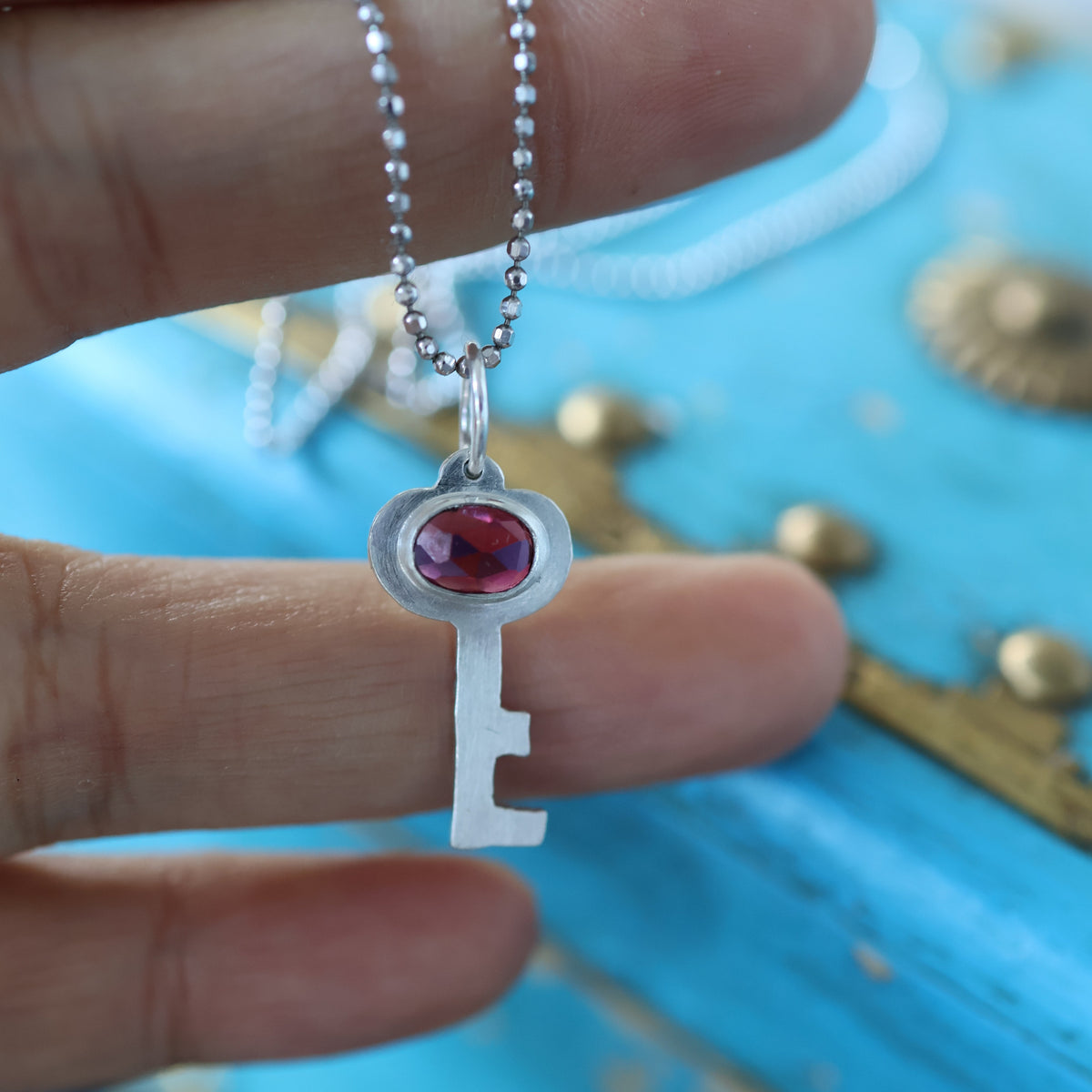 a silver key pendant featuring a red garnet gemstone, on a silver chain held in a hand. in the background is a vintage style blue keepsake box with gold toned hardware.