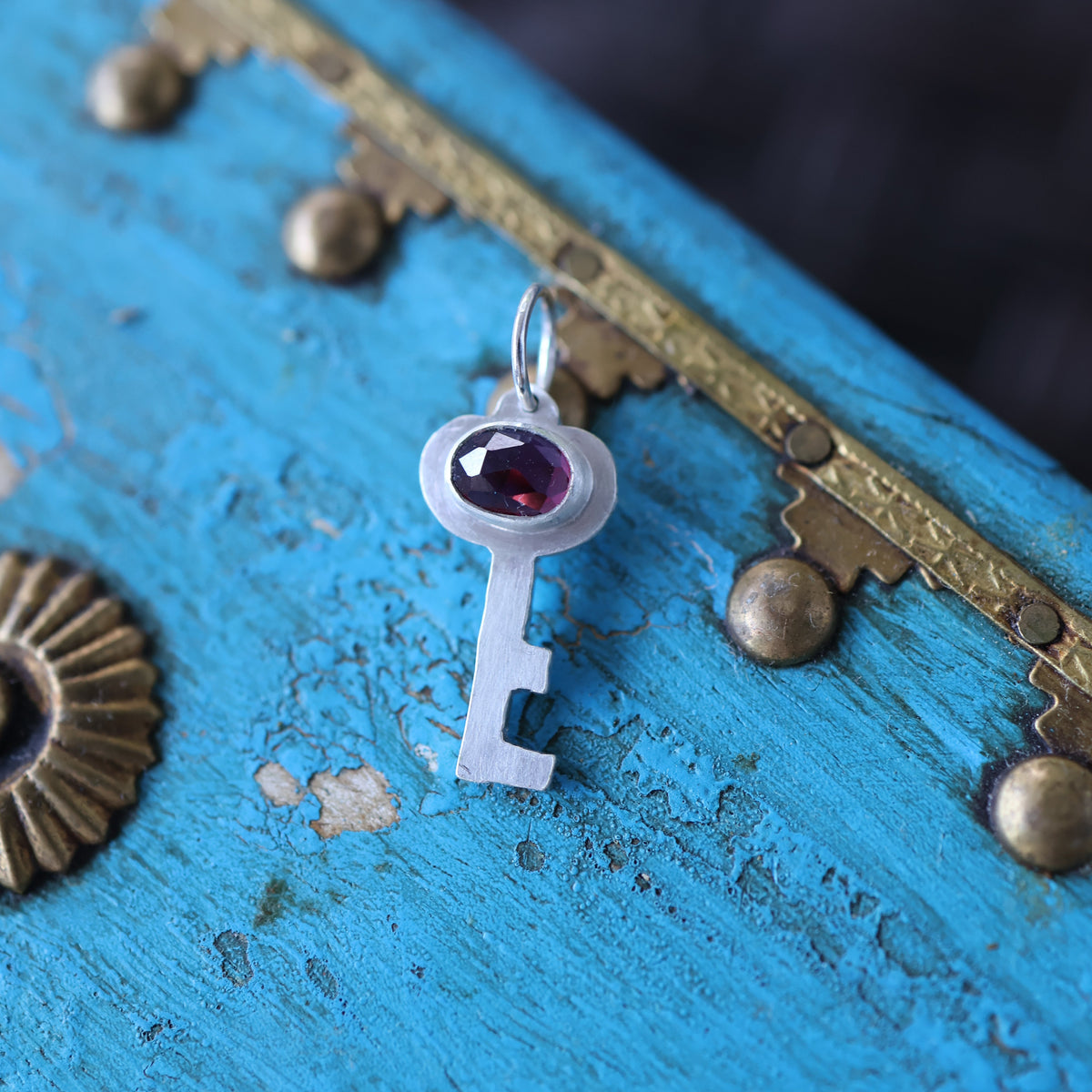 a silver key pendant featuring a red garnet gemstone, on a vintage style blue keepsake box with gold toned hardware.