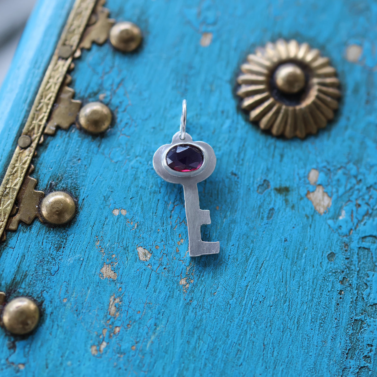 a silver key pendant featuring a red garnet gemstone, on a vintage style blue keepsake box with gold toned hardware.