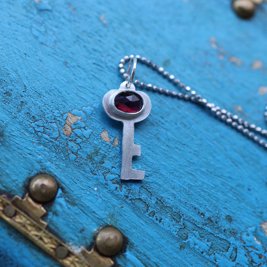 a silver key pendant featuring a red garnet gemstone, on a silver chain. in the background is a vintage style blue keepsake box with gold toned hardware.