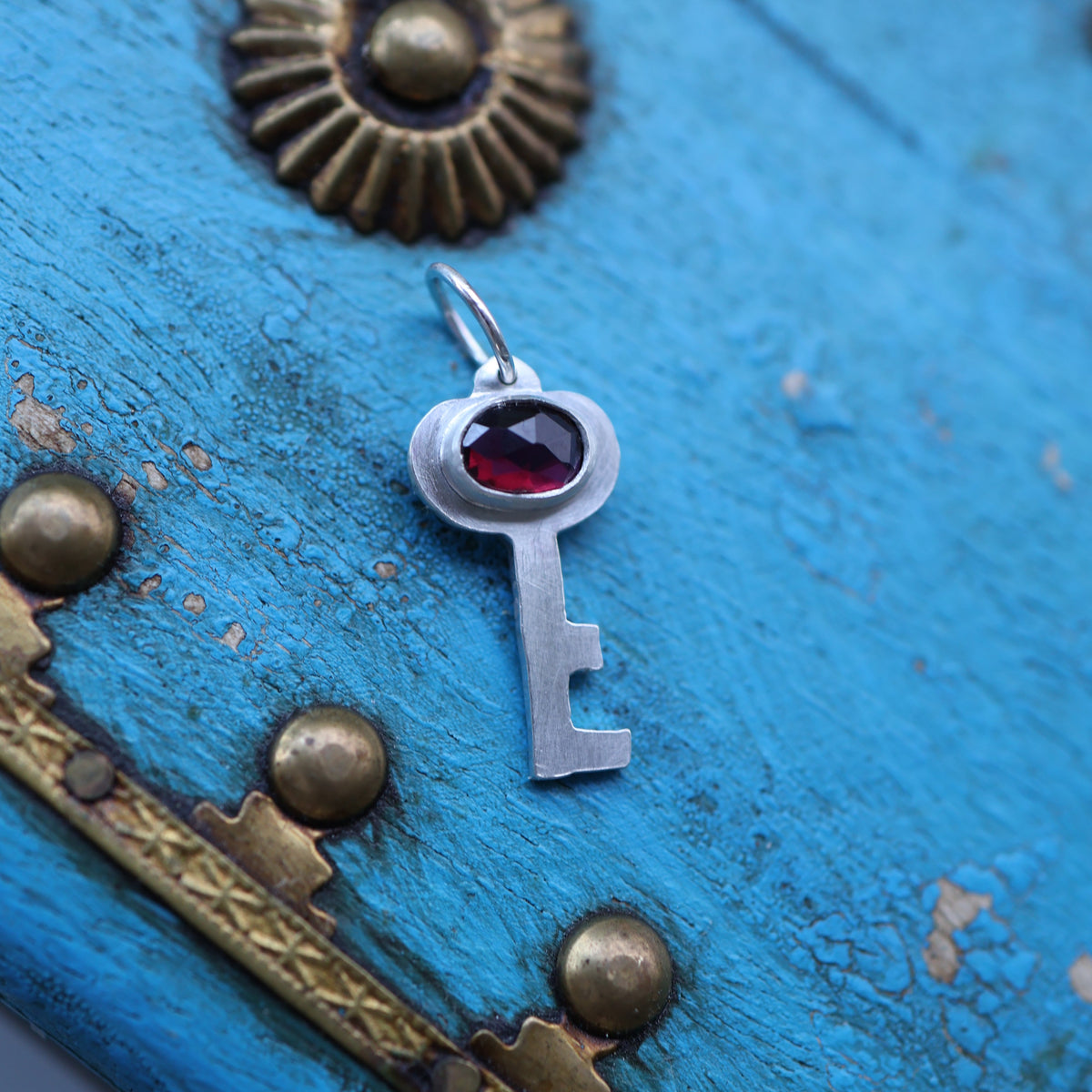 a silver key pendant featuring a red garnet gemstone on a vintage style blue keepsake box with gold toned hardware.