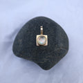 A small gold pendant with a round iridescent moonstone displayed on a dark smooth stone against a pale blue fabric background.