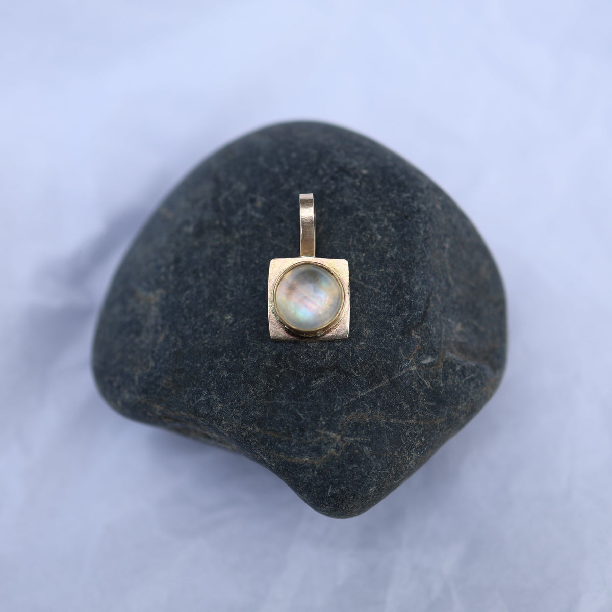 A small gold pendant with a round iridescent moonstone displayed on a dark smooth stone against a pale blue fabric background.