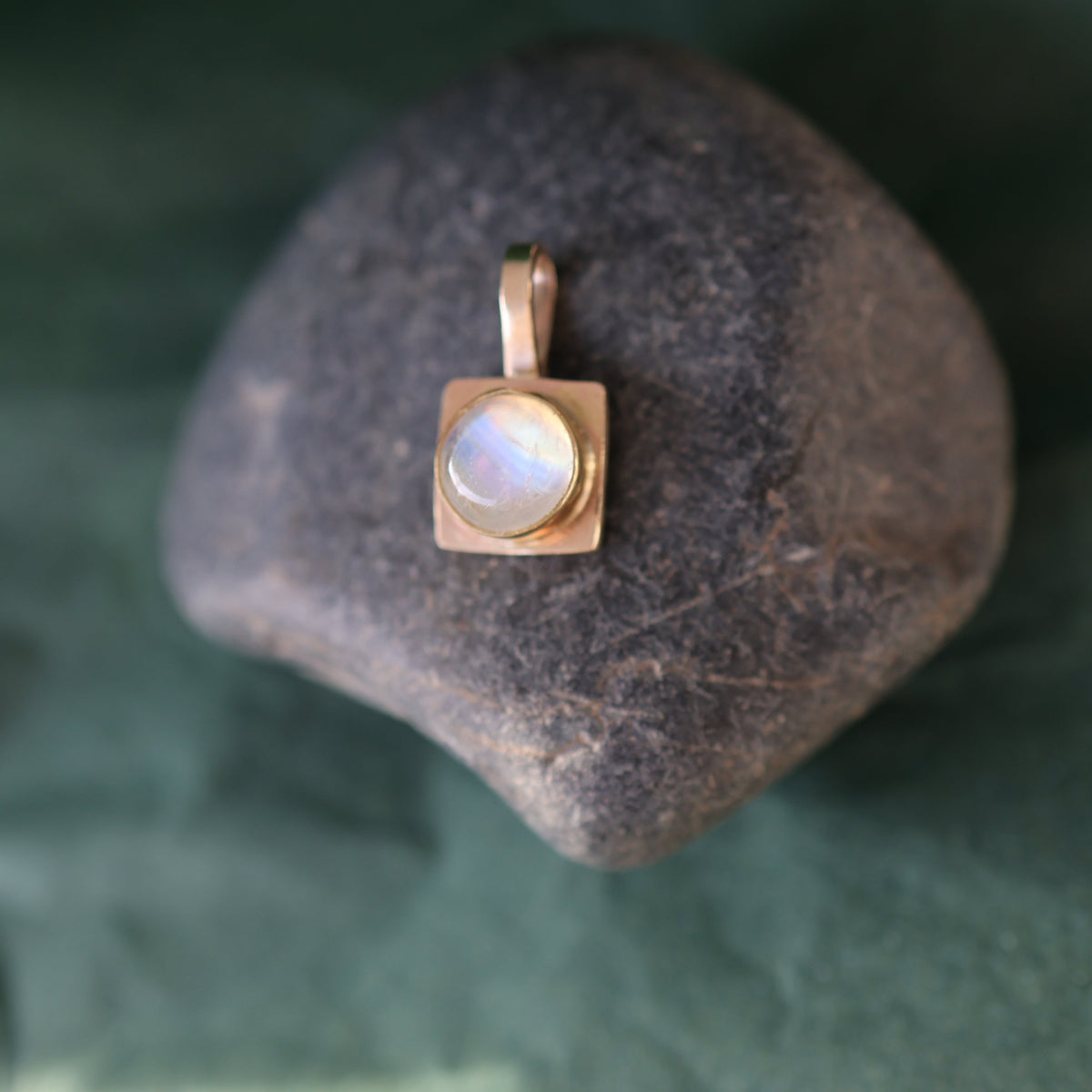 A square gold pendant with a round white moonstone gemstone, displayed on a dark gray stone over a dark green textured fabric background.