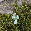 Silver mushroom shaped pendant with a green gemstone on a background of moss and tree bark