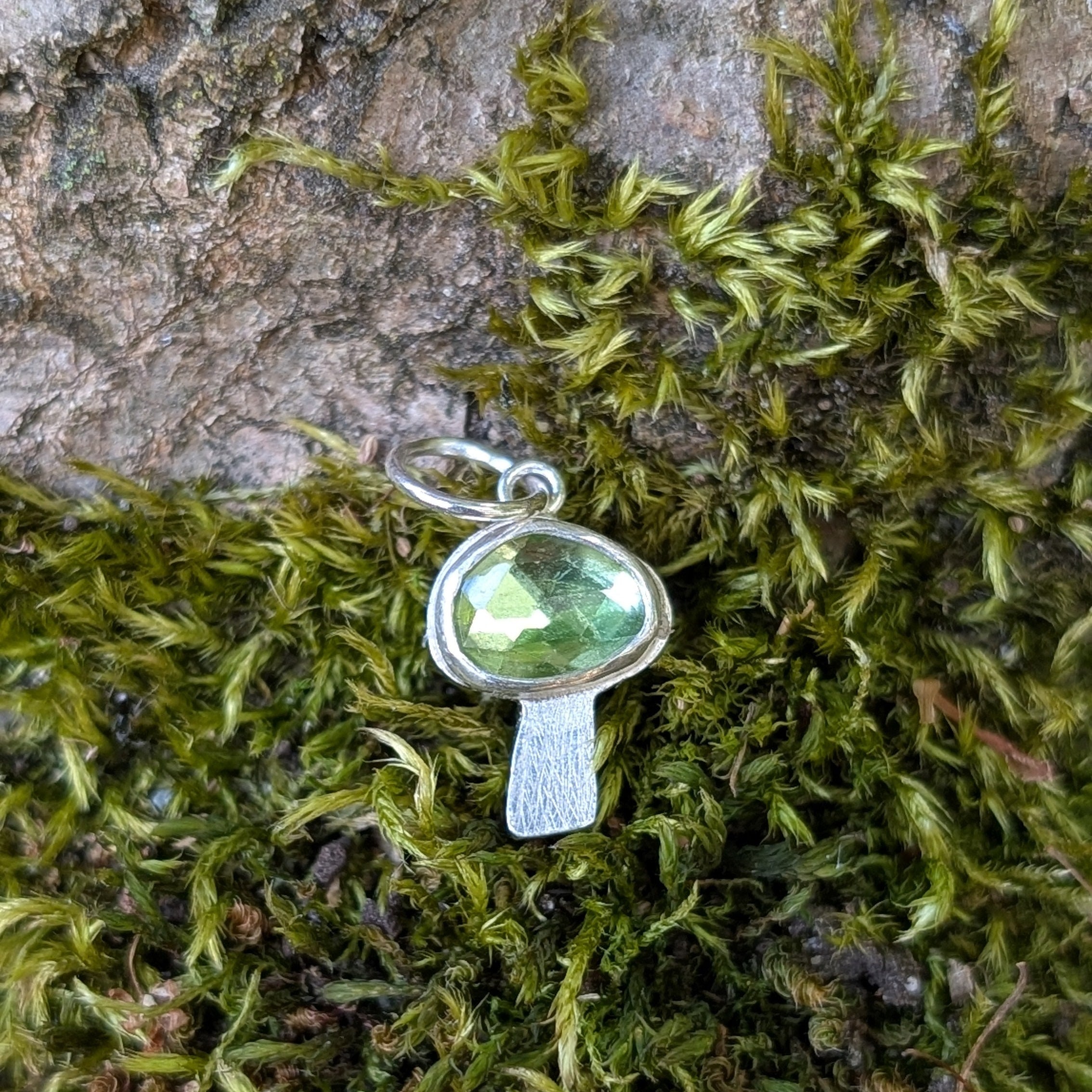Silver mushroom shaped pendant with a green gemstone on a background of moss and tree bark