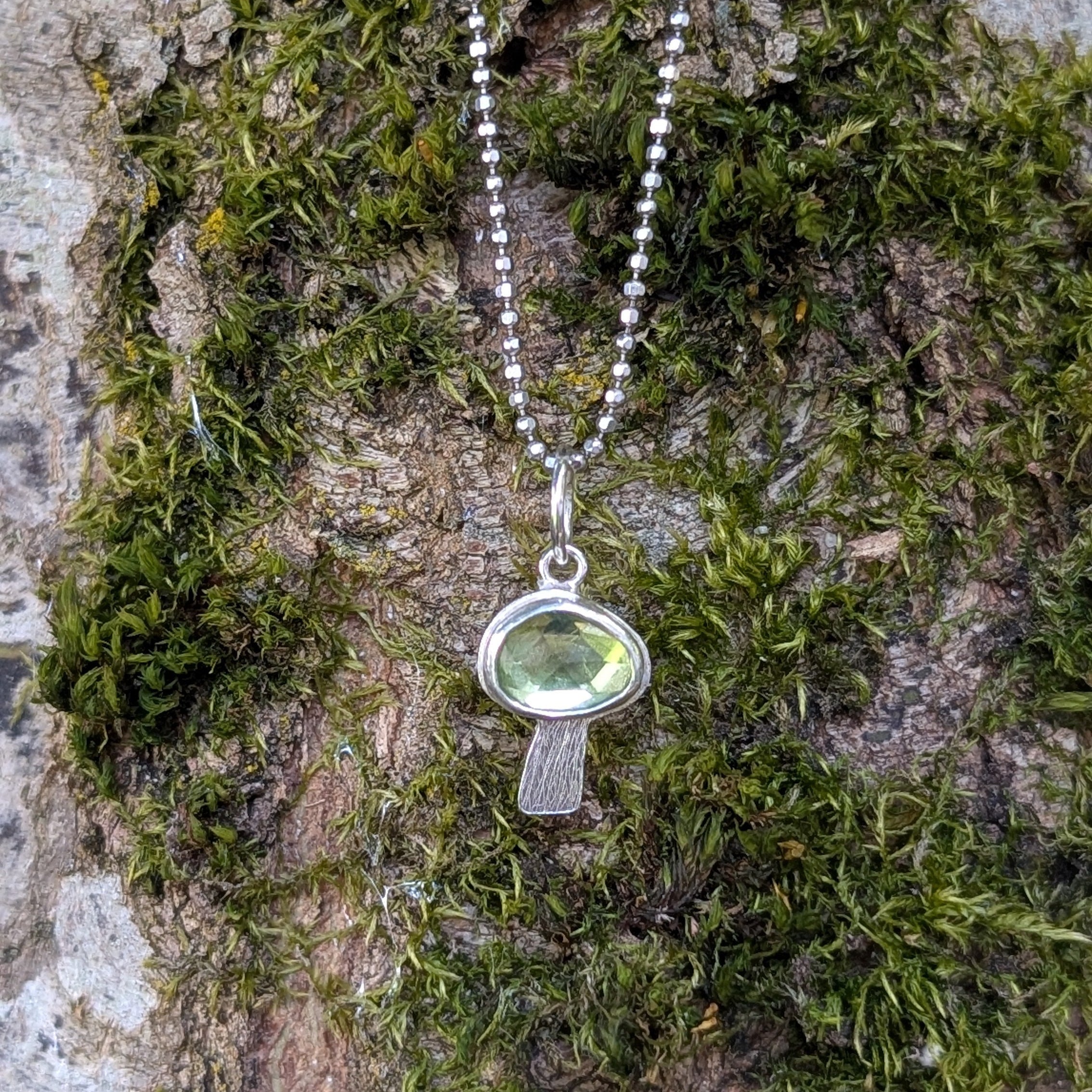 Silver necklace with a peridot mushroom shaped pendant on a background of moss and tree bark