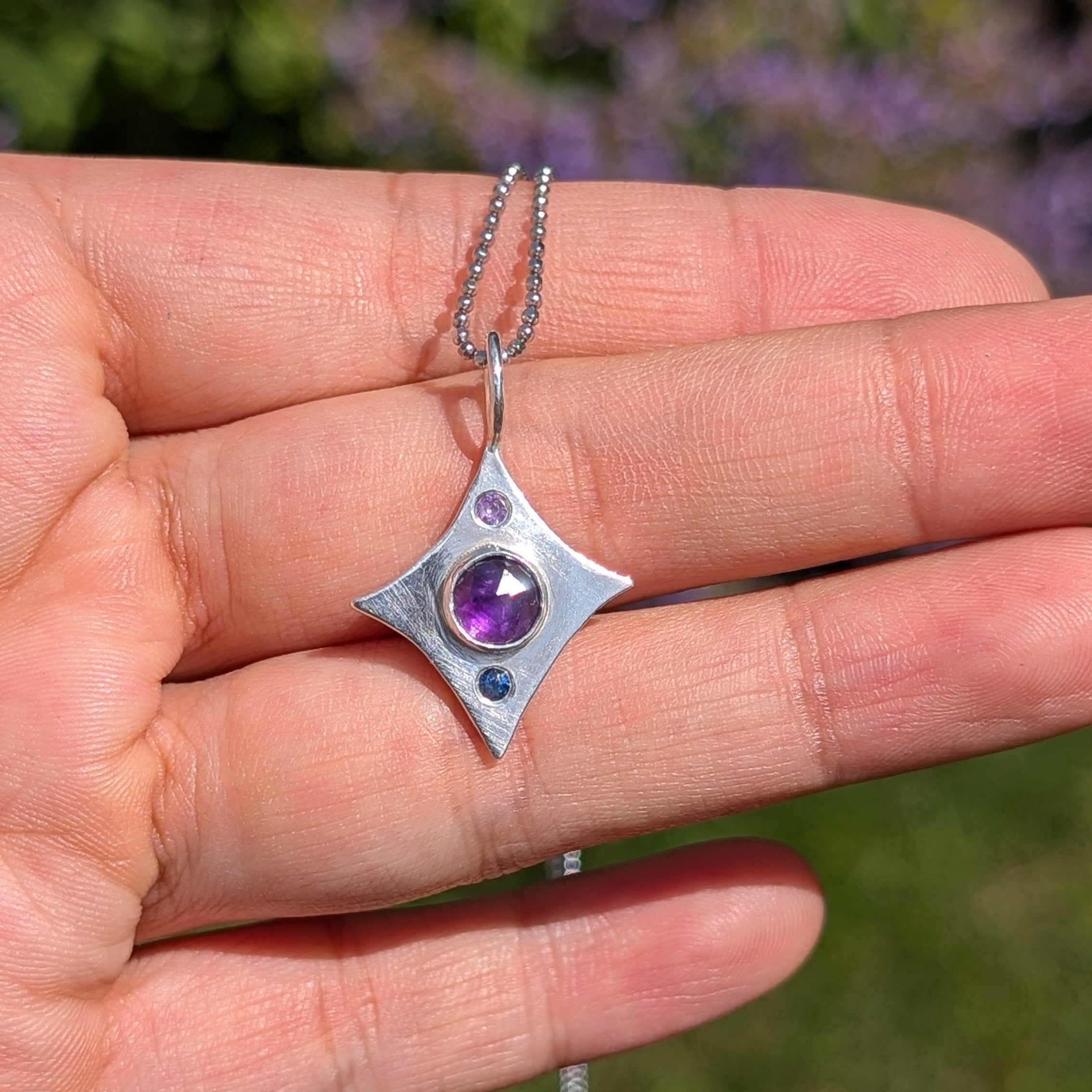 Silver necklace with a pendant featuring a purple gemstone held in a hand against a blurred natural background.