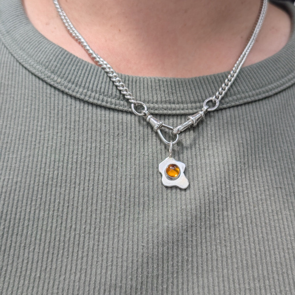 Necklace with a fried egg pendant featuring a colored gemstone on a person wearing a gray shirt.