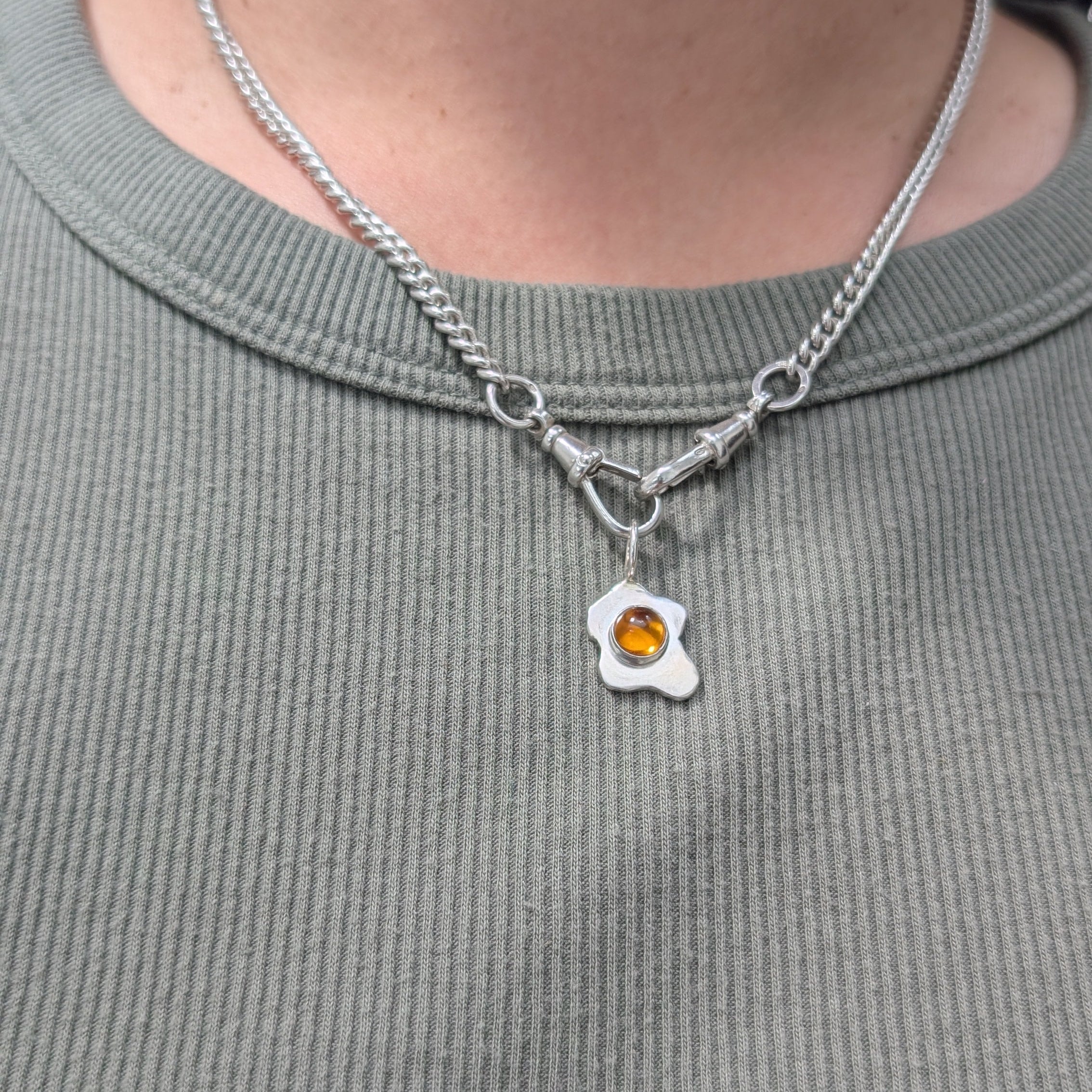 Necklace with a fried egg pendant featuring a colored gemstone on a person wearing a gray shirt.