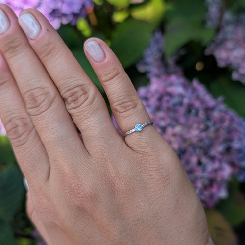 A close-up of a hand wearing a delicate silver ring with a small light blue stone, set against a blurred background of purple hydrangea flowers and green leaves.