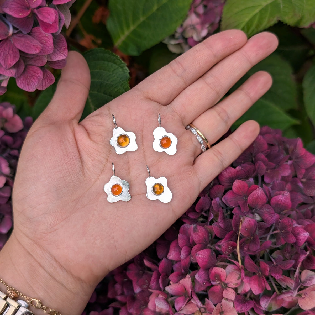 a hand holding four fried egg pendants with orange center gemstones, in front of a floral background