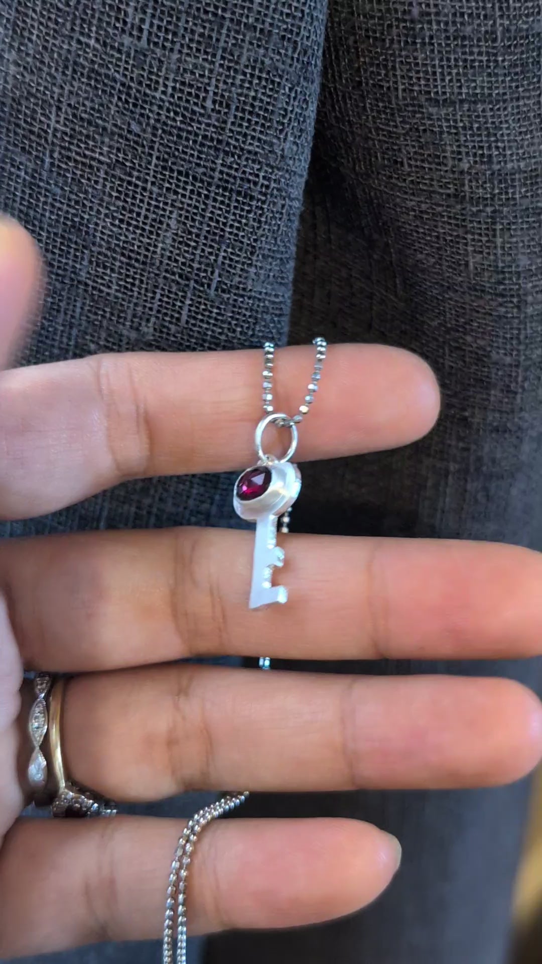 a silver key pendant featuring a red garnet gemstone, on a silver chain held in a hand. 
